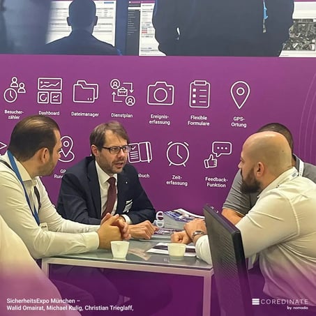 Vier Männer im Gespräch an einem Tisch auf der SicherheitsExpo München, darunter Walid Omairat, Michael Kulig und Christian Trieglaf, vor dem lila COREDINATE®-Messestand mit Symbolen für Besucherliste, Datenmanager, Ereigniserfassung und mehr.