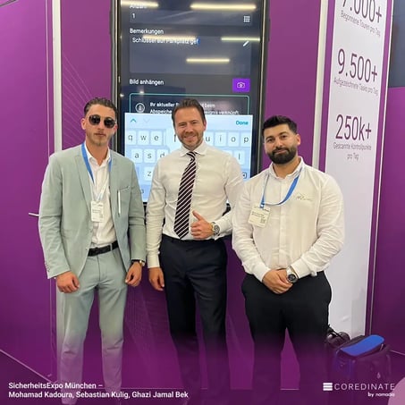 Mohamad Kadoura, Sebastian Kulig und Ghazi Jamal Bek stehen nebeneinander und lächeln in die Kamera am COREDINATE®-Messestand auf der SicherheitsExpo München.
